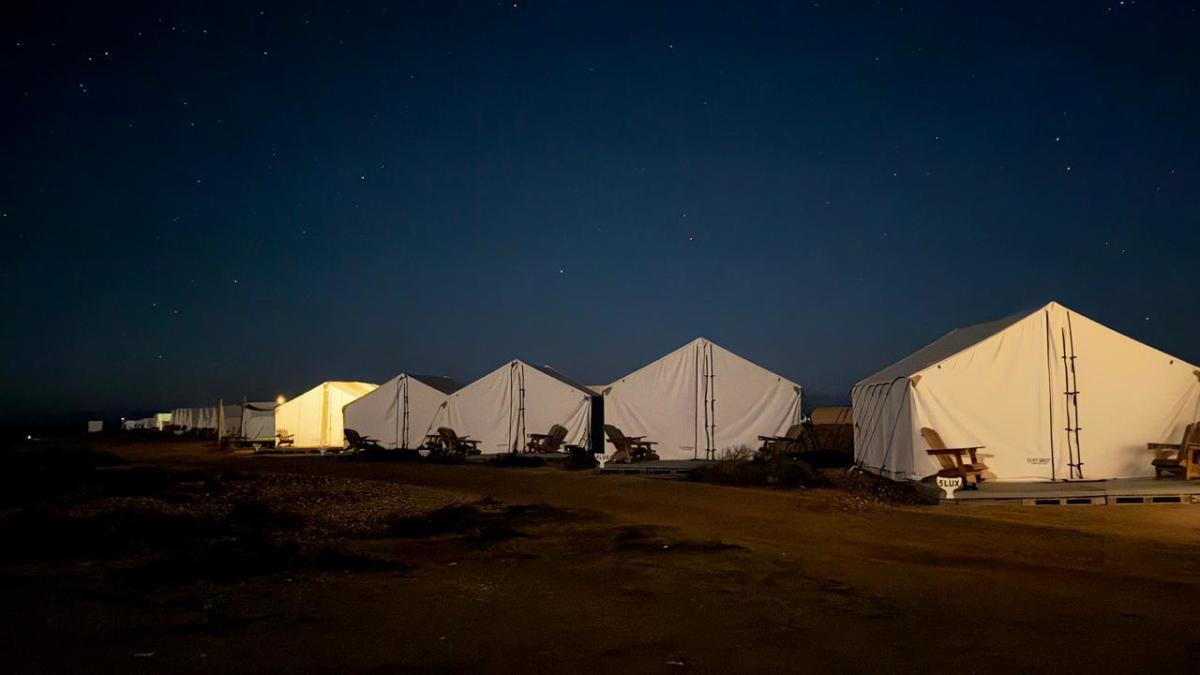 A row of large white glamping tents pitched on wooden platforms under a clear, starry night sky. One tent glows with warm yellow light from within, and wooden chairs sit outside each tent on the dark ground.