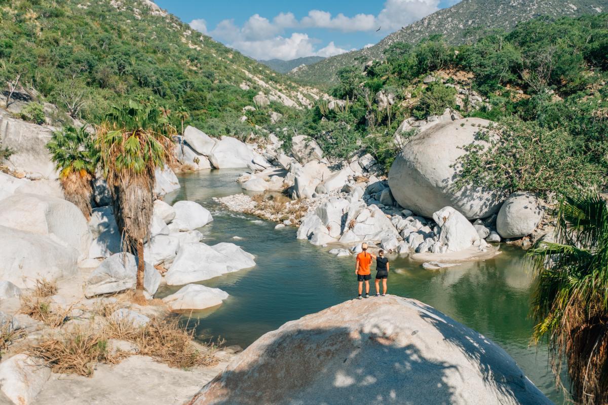 Two hikers stand atop a massive boulder overlooking a freshwater pool and palm-lined canyon in the Sierra de la Laguna mountains.