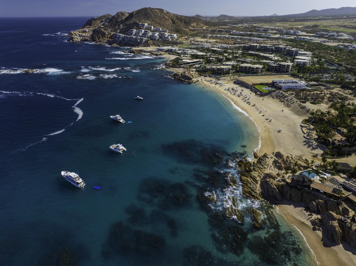 Aerial view of the crescent-shaped Chileno Bay in Los Cabos, featuring vibrant turquoise water, white yachts at anchor, and a golden sand beach lined with luxury resorts and rugged desert hills under a clear blue sky.