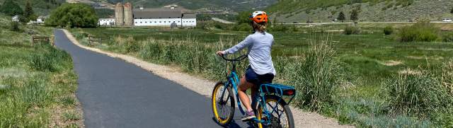 Woman riding an electric bike along a paved path with a white barn in the background