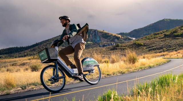 A rider on a Summit Bike Share e-bike rides on a paved path in front of the Utah Olympic Park in Park City, UT