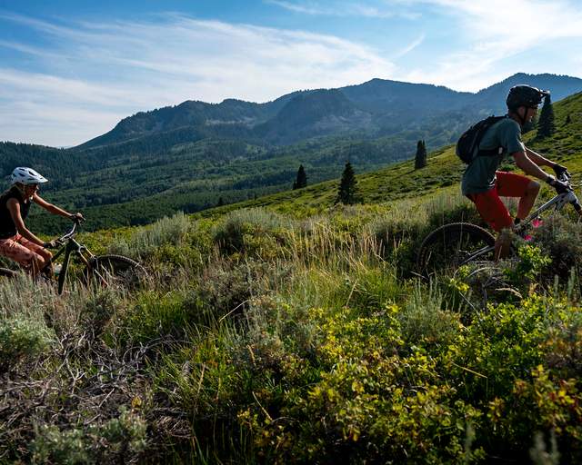 A father and daughter ride on a trail surrounded by green foliage and mountains on a sunny day in Park City, UT
