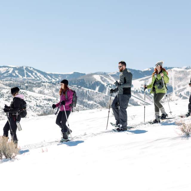 Group of six adults snowshoeing on a sunny day with ski resort in background