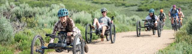 Adaptive mountain bikers ride up a wide dirt path on a green hill in Park City, UT