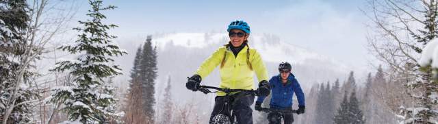 Two people in a yellow and blue jacket mountain biking through the snow under a clear sky.