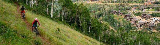Two mountain bikers ride a trail surrounded by green grass overlooking Deer Valley in Park City, UT