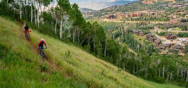 Two mountain bikers ride a trail surrounded by green grass overlooking Deer Valley in Park City, UT