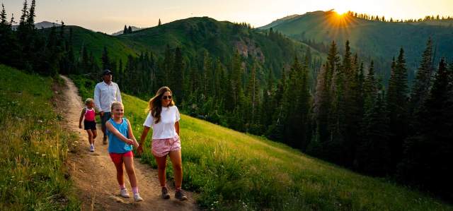 A family of four hikes on a trail above a green forest at sunset in Park City, UT