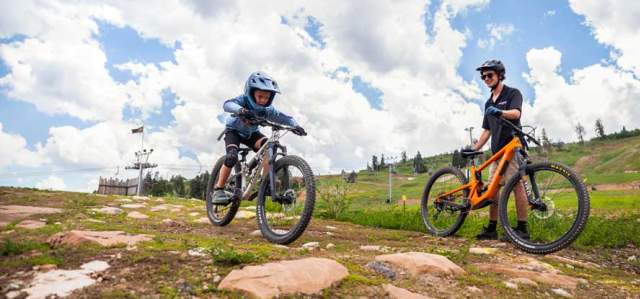 A client rides their mountain bike over a rock feature while a coach looks on at Woodward in Park City UT