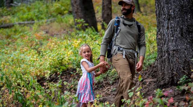 Alpine Forestry owner, David Telian, hikes with his young daughter on a trail surrounded by low green vegetation and tall trees near Park City, UT