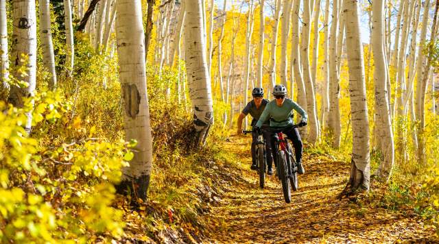 Two riders smile as they ride their bike through fall leaves on a trail near Park City, UT