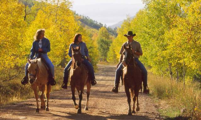 Three people ride horses down a dirt road surrounded by fall foliage in Park City, UT