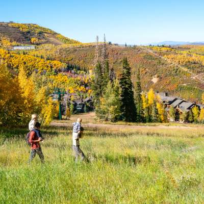 Two parents hike with their children riding on shoulders on a trail in front of Fall leaves in Park City, UT