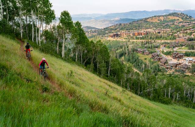 Two mountain bikers ride a trail surrounded by green grass overlooking Deer Valley in Park City, UT