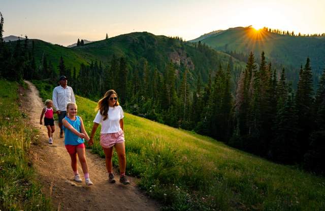 A family of four hikes on a trail above a green forest at sunset in Park City, UT