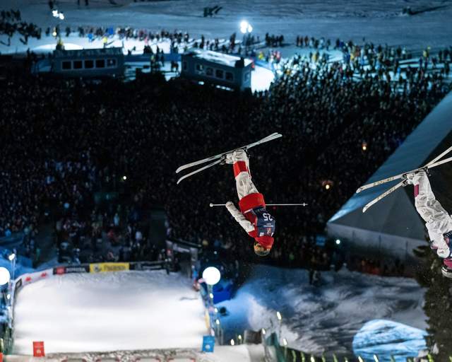 Two skiers perform aerial tricks at night on a lit course above a crowd at the Deer Valley World Cup in Park City, UT