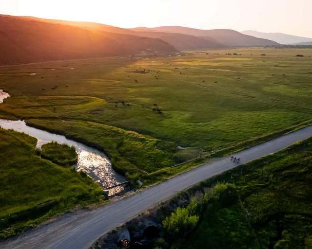 Three cyclists ride on a gravel road crossing a creek at sunset surrounded by green fields in Summit County, UT.