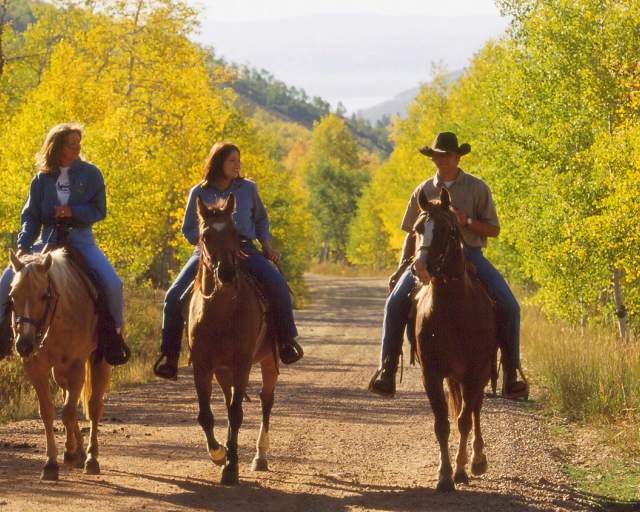 Three people ride horses down a dirt road surrounded by fall foliage in Park City, UT