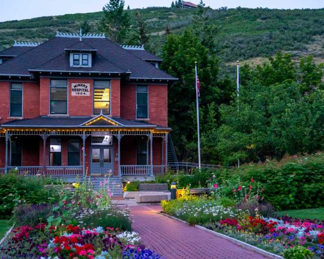 The Old Miners Hospital surrounded by lush green trees and bright flowers at dusk in Park City, UT