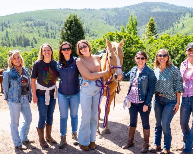 A group of women pose for a photo with a light brown horse with green hills in the background in Park City, UT