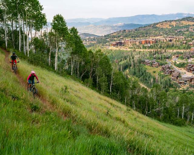 Two mountain bikers ride a trail surrounded by green grass overlooking Deer Valley in Park City, UT