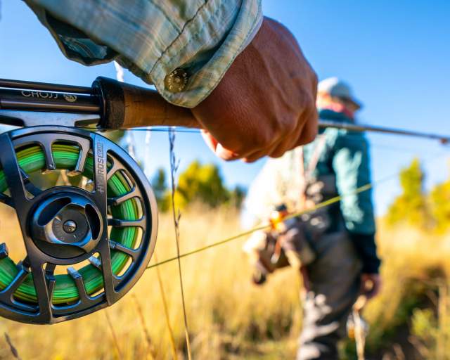 A hand holds a fly rod and reel in a dry grass field on a sunny day in Park City, UT