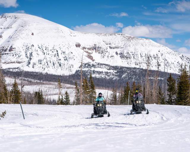 Two snowmobilers ride up a road in front of snow covered mountains and forest in the Uinta Mountain Range near Park City, UT