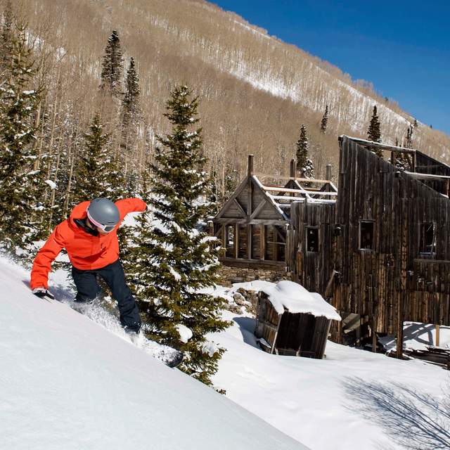 A snowboarder carves a turn across a run in front of a historic mine building on a sunny day at Park City Mountain