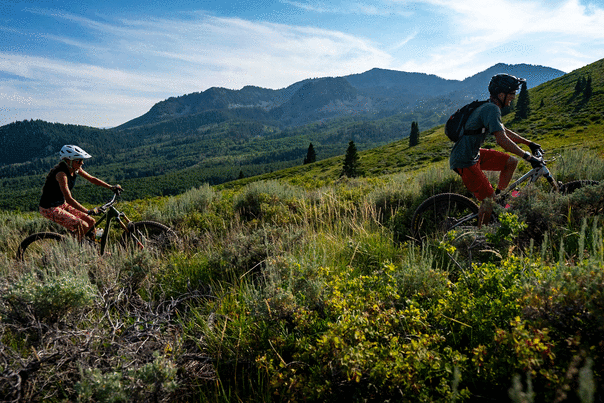 A father and daughter ride on a trail surrounded by green foliage and mountains on a sunny day in Park City, UT