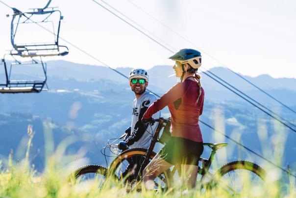 Two mountain bikers smile on and look at a view with a chair lift in the background in Park City, UT