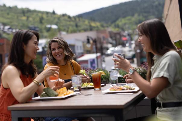 Three women laugh and talk together while eating a meal on a rooftop overlooking Historic Main Street, Park City, UT.