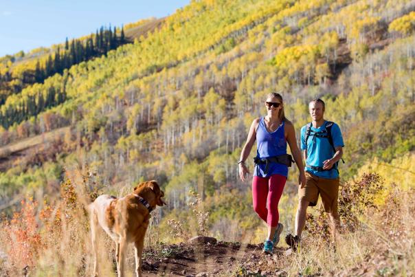 A couple and a dog hike on a trail with fall foliage in the background in Park City, UT