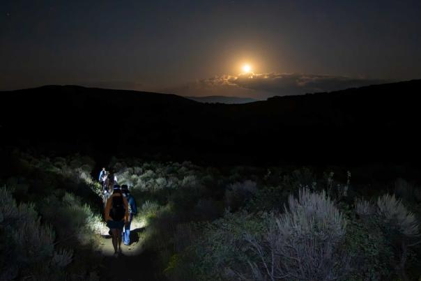 A group of hikers walk down a dark hill lit by head lamps with a full moon over a band of clouds in Round Valley in Park City, UT