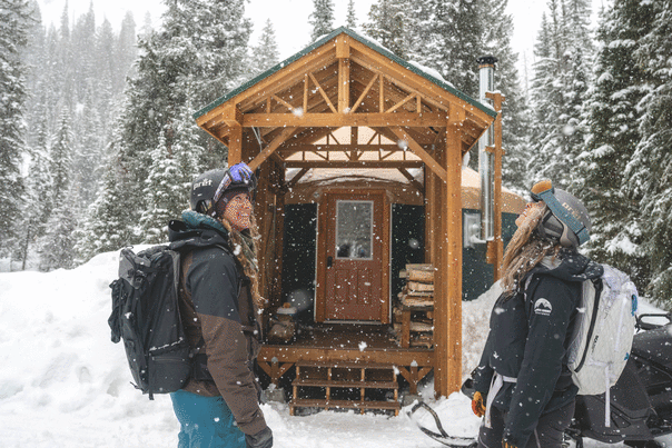 Two skiers look up at falling snow in front of a snow covered yurt in the woods in the Uinta Mountains near Park City, UT