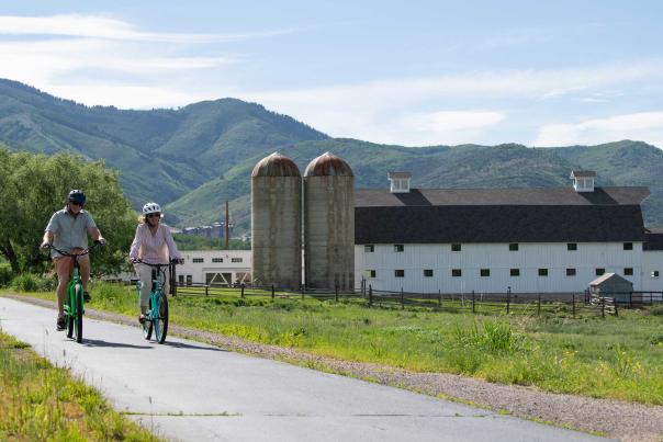 Two people biking a paved trail with the white McPolin Farm in the background.