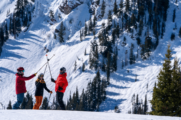 Three skiers raise their ski poles in front of steep terrain on sunny day at Deer Valley in Park City, UT