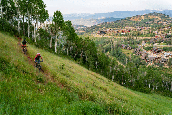 Two mountain bikers ride a trail surrounded by green grass overlooking Deer Valley in Park City, UT