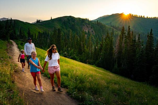 A family of four hikes on a trail above a green forest at sunset in Park City, UT