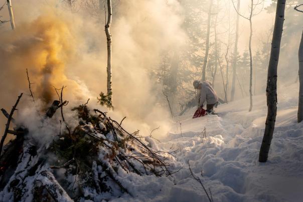 A worker shovels snow toward a prescribed burn pile while surrounded by smoke in a forest near Park City,  UT