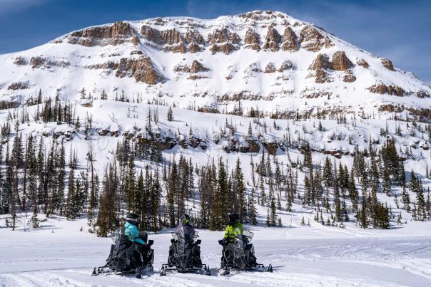 Three people talk while sitting on snowmobiles in front of a large snow covered mountains in the Uintas near Park City, UT
