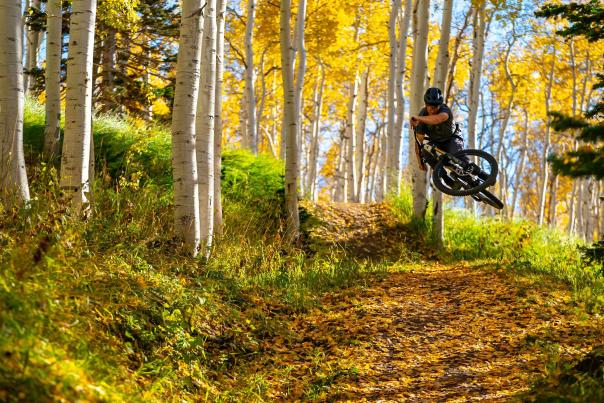 A mountain biker tables a gap jump on a trail covered in yellow fall leaves in Park City, UT