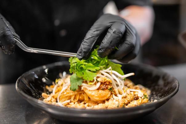 A gloved pair of hands plates a serving of Pad Thai in a kitchen in Park City, UT