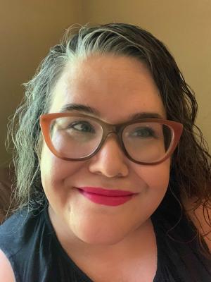 Close-up photograph of Johannah Maynard Edwards, a smiling white woman with light-medium skin tone. Her hair is salt and pepper, wavy and parted at the side. She is wearing brown cat-eye shaped glasses over her blue eyes which are looking to the right of the camera. She wears a fuschia pink lip tint.