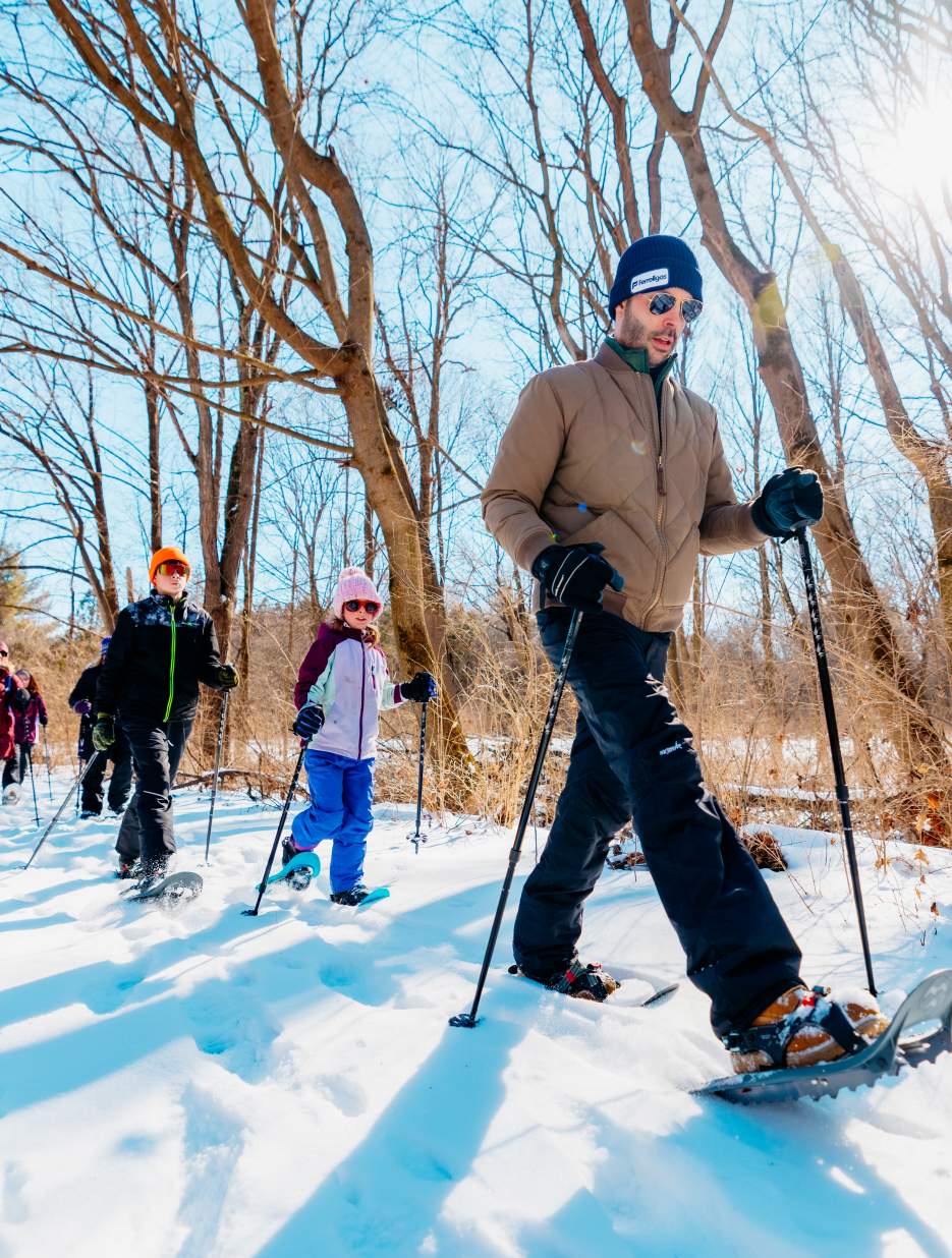 A group of people snowshoeing on a snowy trail at Rock Cut State Park in Rockford, IL, surrounded by bare trees under a bright blue sky.