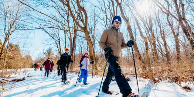 A group of people snowshoeing on a snowy trail at Rock Cut State Park in Rockford, IL, surrounded by bare trees under a bright blue sky.