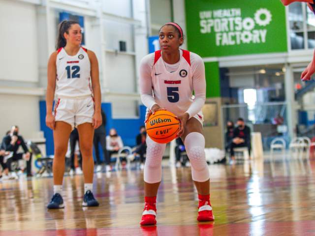 Two female basketball players, one holding a basketball on a polished court, with spectators and a "Sports Factory" sign in the background.