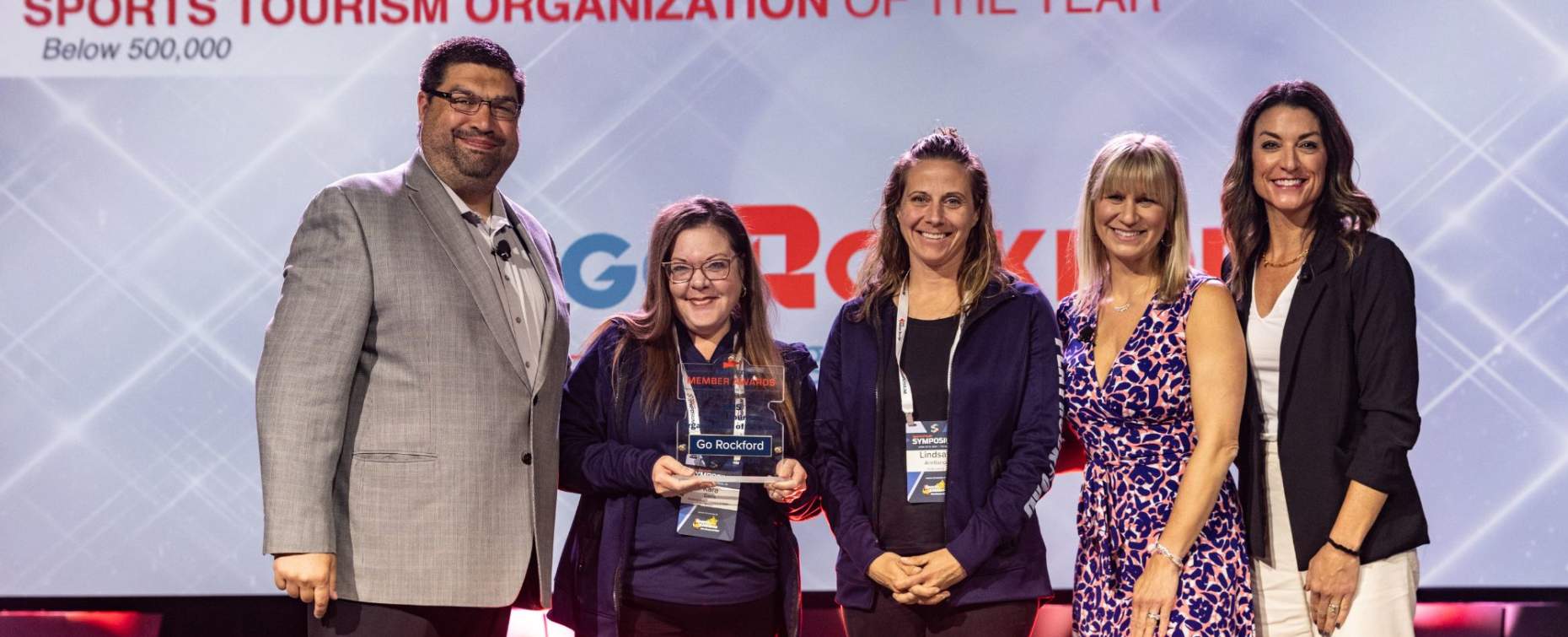 Group of award recipients standing on stage and holding a trophy during the Sports ETA Sports Tourism Organization of the Year award presentation.