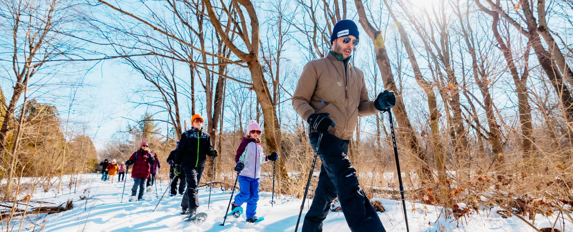 A group of people snowshoeing on a snowy trail at Rock Cut State Park in Rockford, IL, surrounded by bare trees under a bright blue sky.