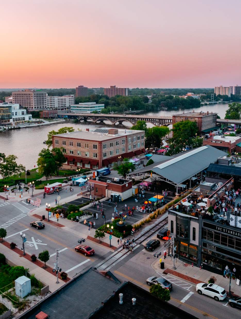 Aerial view of downtown Rockford at sunset, with people gathering near food trucks along a lively riverside, surrounded by city buildings.