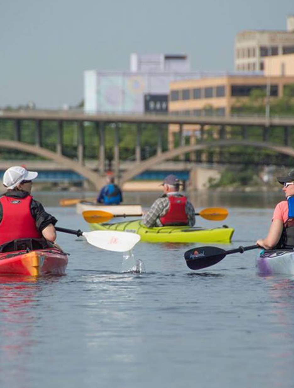 Kayakers paddling on a Rockford, IL calm river, with a bridge in the background and city buildings along the shore.